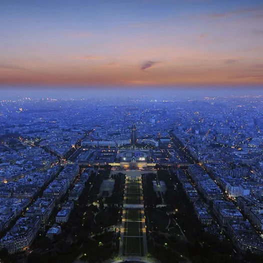 Aerial view of Paris cityscape at twilight, with illuminated buildings and streets stretching to the horizon, taken from high above showing the Trocadéro gardens and city lights below a gradient pu...