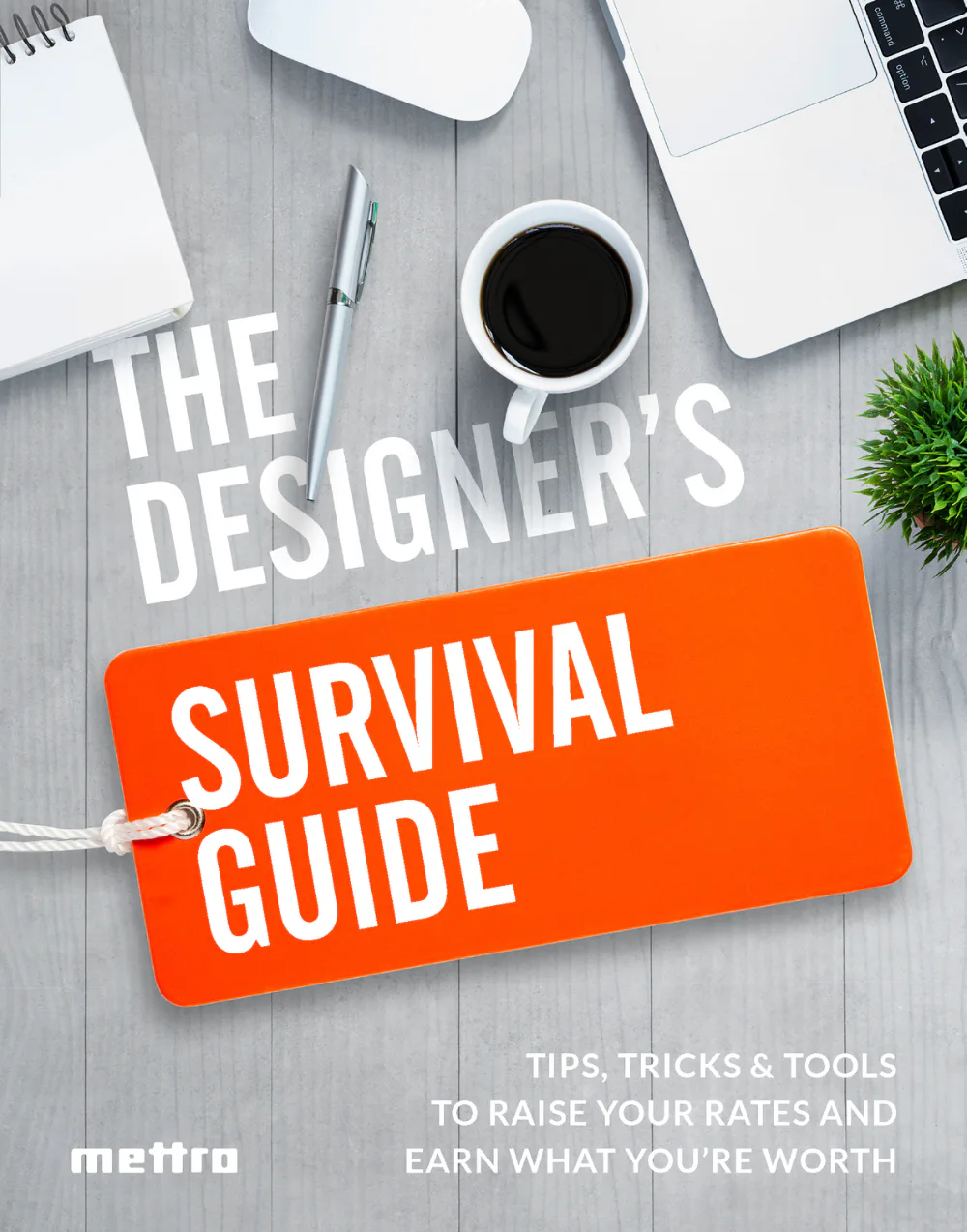 Flat lay workspace showing 'The Designer's Survival Guide' orange tag with white text on gray wooden desk, surrounded by laptop, coffee cup, notepad, pen, wireless mouse, and green plant.