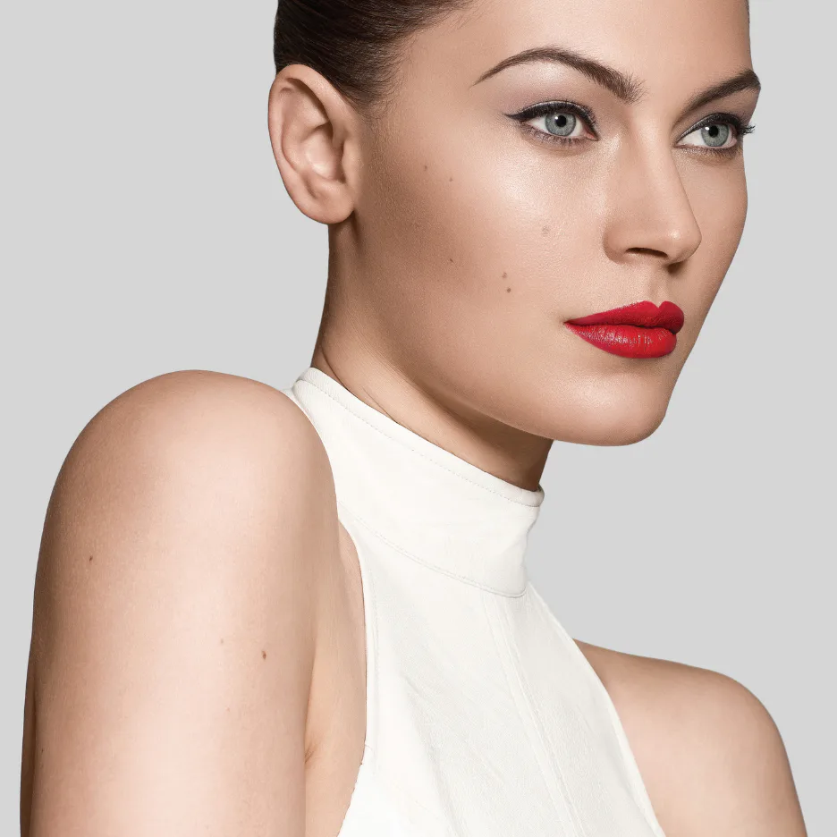 Woman with flawless skin displaying French beauty makeup featuring bold red lips, defined eyebrows, and neutral eyeshadow, wearing white halter top against neutral background