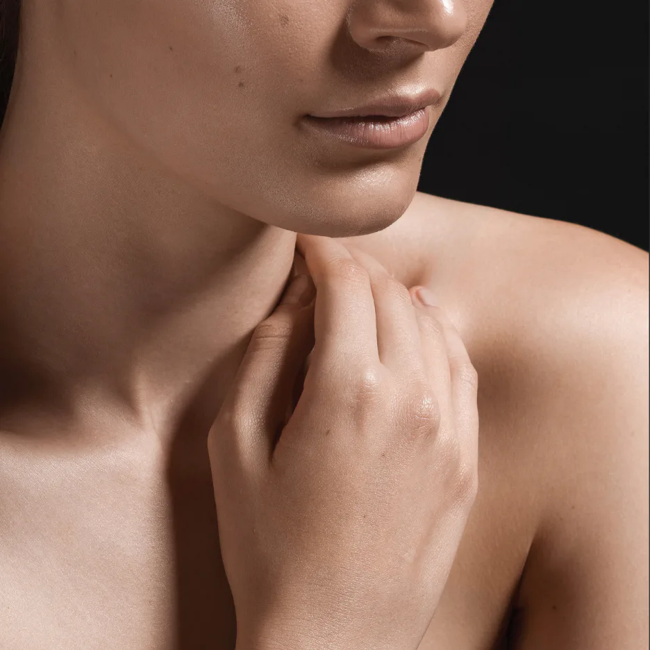 Close-up of a person's lower face and neck with glowing, moisturized skin. Fingers gently touch the chin and neck area, demonstrating skincare technique and skin texture.