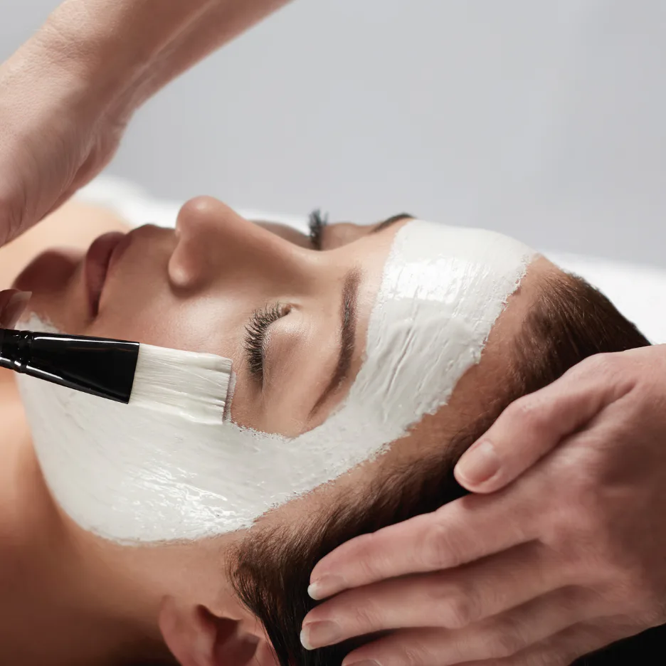 Aesthetician applying white facial mask to client's face with brush while client relaxes with eyes closed, wearing white headband during spa treatment