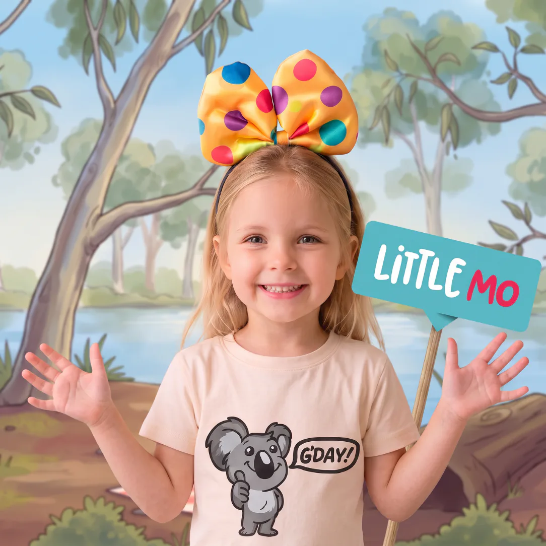 Smiling young girl wearing cream t-shirt with koala graphic, colorful polka-dot bow headband, and holding 'Little Mo' sign against illustrated forest background with trees and water